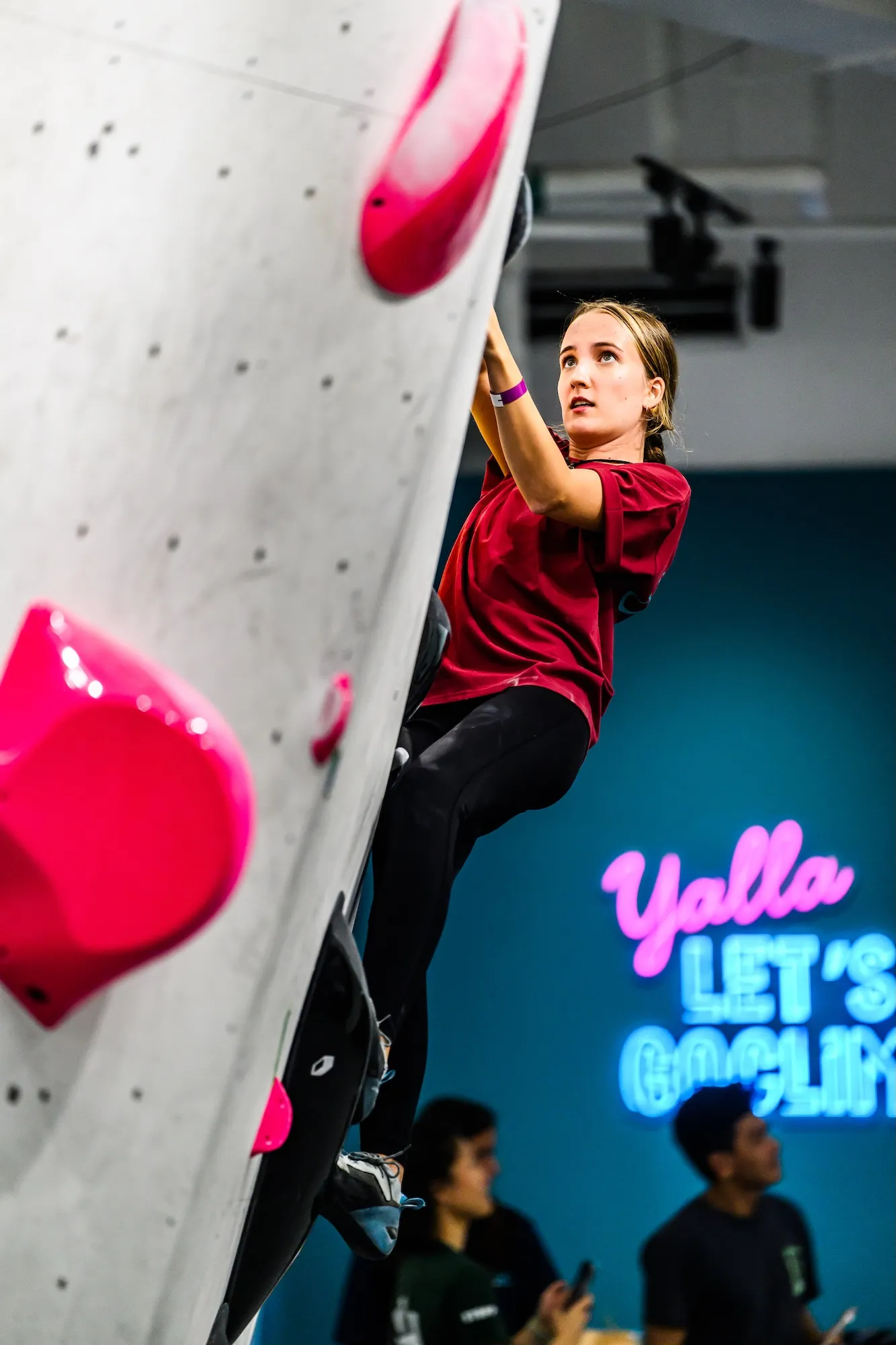 A climber working a steep line above the crowd, the gym's neon sign glowing behind
