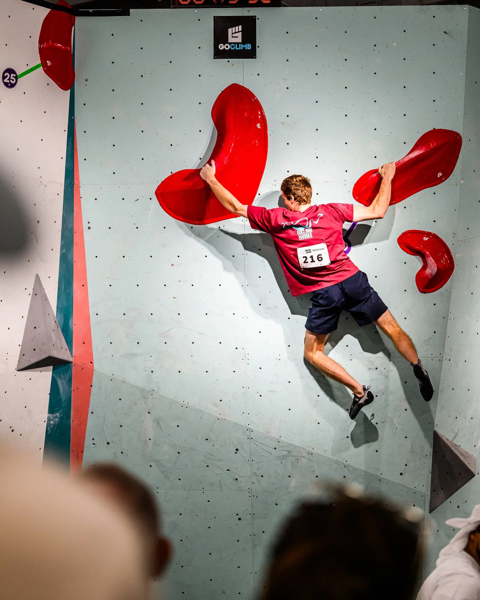 A competitor in a numbered bib spanning between two large red holds, watched from the floor