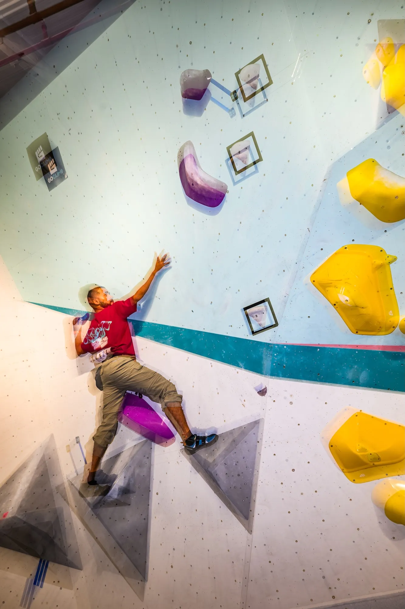 A competitor reaching high on the teal competition wall, one foot planted on a purple volume